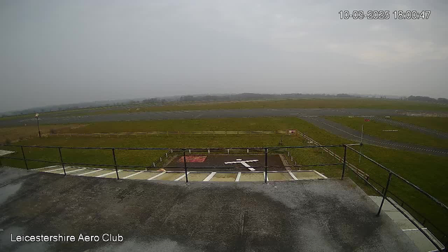 A view from a webcam at Leicestershire Aero Club showing a grassy airfield under a cloudy sky. In the foreground, there is a concrete area with a large white cross marking, indicating a helipad. Beyond that, a runway stretches across the right side of the image, with grassy fields and trees in the distance. There is a railing along the edge of the viewing platform and a small red sign visible near the runway. The overall scene appears overcast and somewhat muted in color.