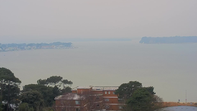 A hazy view of a calm body of water with distant land visible on the horizon. In the foreground, there is a brown, multi-story building partially obscured by trees. The sky is grey and overcast, blending with the muted colors of the water. There are small boats near the shoreline on the left side of the image, and the area in the background appears to have some structures, likely part of a coastal community.