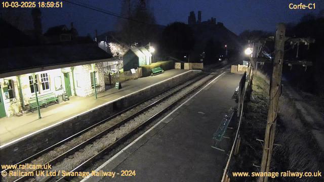 A railway station platform is viewed at dusk, with soft lighting illuminating the scene. To the left, there is a station building with stone walls, large windows, and green accents. Several green benches are placed along the platform. A sign marked "WAY OUT" is visible. The platform is lined with tracks, and a wooden fence can be seen in the distance. In the background, the silhouettes of hills and structures are faintly visible against the darkening sky. A lamp posts illuminates part of the scene, contributing to the evening ambiance.