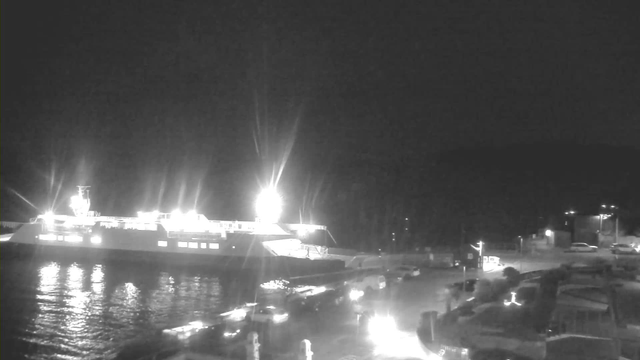 A brightly lit ferry docked at a dark harbor. The ferry has strong lights illuminating the area, reflecting off the water. Several vehicles are parked along the dock, and some faint lights are visible in the background, indicating nearby structures. The scene appears to be at night, with dark surroundings contrasting with the bright lights of the ferry and vehicles.