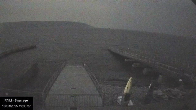 A dimly lit scene of a shoreline with a long jetty extending into the water. The calm sea reflects minimal light, and there are outlines of hills or land in the background. In the foreground, a concrete surface is visible with safety barriers, and several small boats or kayaks are stored near the edge.