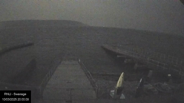 A dark, grainy image captured from a webcam overlooking the sea. The scene features a partially visible jetty that extends into the water, with a small kayak or canoe on the dock, angled to the right. The water looks choppy under a cloudy sky. In the background, a hill rises above the shoreline, fading into the muted light of dusk. A timestamp is located at the bottom left corner, indicating the date and time of the image.