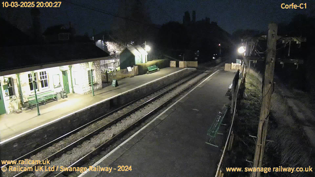 A dimly lit railway station at night is captured in the image. The platform has a long, straight section with two metal tracks running parallel to it. To the left, there is a stone building with multiple windows, some of which are illuminated. Green benches are visible on the platform along with a sign that reads "WAY OUT." On the right side, there is a wooden fence with some greenery, and a light post is visible, casting a soft glow. The scene is quiet and devoid of people or trains at this moment.