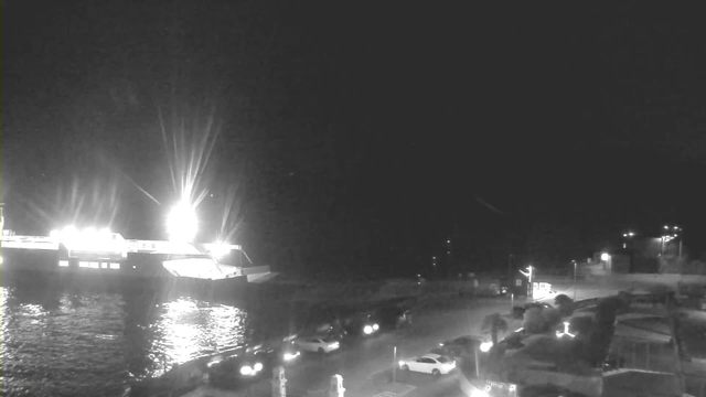 A nighttime view of a harbor scene. In the foreground, several parked cars are visible along the road. To the left, a large ferry is illuminated by bright lights as it prepares to dock, casting reflections on the water. The shoreline has faint outlines of buildings and streetlights, contributing to the overall dark atmosphere of the image. The background is mostly obscured by darkness.