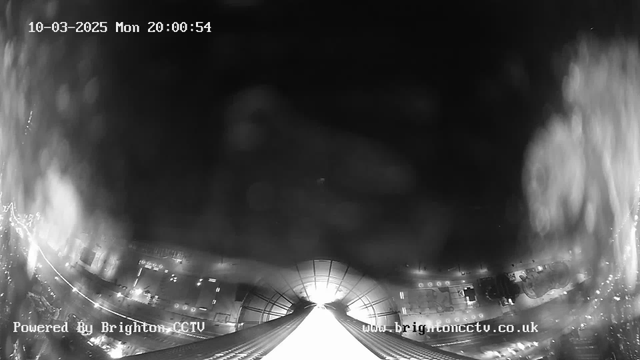 A black and white image captured from a webcam, showing a view down a tall structure, likely a tower or building. The top of the image features a bright light source, possibly indicating a light shining from above or an opening. In the lower part of the image, faint outlines of streetlights and roads are visible, suggesting there is urban infrastructure below. Some splatters or reflections indicate moisture on the camera lens. The timestamp reads 10-03-2025 at 20:00:54. The bottom left corner includes text that says "Powered By Brighton CCTV" and the website URL visible at the bottom right.