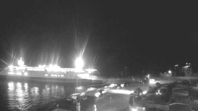 A ferry docked at a harbor at night, illuminated by bright lights. The water reflects the light, creating a shimmering effect. In the foreground, vehicles are parked on the quay, with the path lit by headlights. The scene is mostly dark, with some distant lights visible along the shoreline.