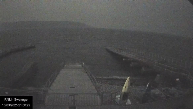 A dark, hazy scene showing a dock extending into a body of water. The area appears to be tranquil, with low waves. A small section of the dock is visible, along with two kayaks or canoes in muted colors, one yellow and one dark. The background features vague contours of hills or land, shrouded in darkness. The scene has a serene but eerie atmosphere, suggesting night-time conditions. A timestamp indicates the date as March 10, 2025, at 21:00:29.