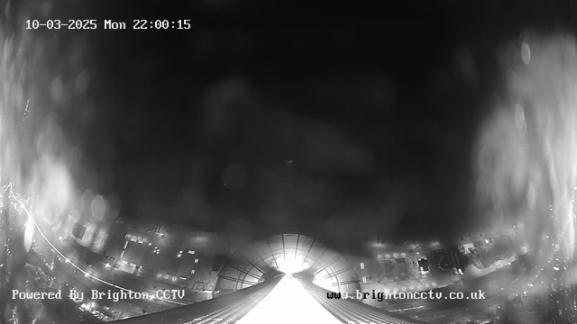 A nighttime webcam image features a wide view of a brightly lit area in the foreground, with blurred lights and shapes suggesting buildings or vehicles. The image is predominantly dark, with a bright white light source at the center, which may indicate an entrance or exit. The timestamp in the top left corner reads "10-03-2025 Mon 22:00:15." The bottom left corner displays the text "Powered By Brighton CCTV," and the bottom right shows "www.brightoncctv.co.uk."