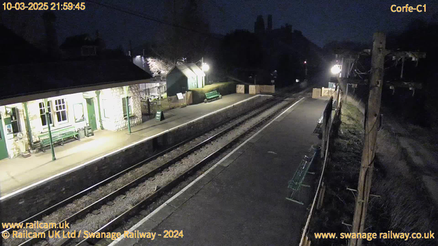 A dimly lit railway station at night. On the left, there's a stone building with green detailing, likely a waiting area, with benches in front. The platform is empty, and there are two parallel railway tracks visible leading into the darkness. A sign that reads "WAY OUT" is positioned at the end of the platform. Several wooden fences and a green seat are present along the platform. In the background, faint lights illuminate the area, suggesting nearby buildings and trees.