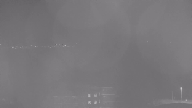 A monochrome image of a dimly lit scene showing faint outlines of buildings and lights in the distance. The view appears hazy, with a foggy quality that obscures much of the detail. There are several small points of light that suggest a distant urban area, and the foreground contains indistinct shapes that could be structures or trees.