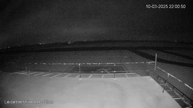 A dark and dimly lit scene viewed from a webcam, showing a flat surface with faint outlines of a helipad. The foreground features a railing, creating a boundary. The background is mostly dark, with distant lights barely visible on the horizon. The time and date are displayed in the upper right corner, reading 10-03-2025 at 22:00:50. The view is tranquil and quiet, with no obvious activity visible. The scene lacks color and is primarily monochromatic.