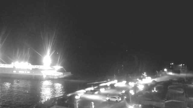 A nighttime scene featuring a large ferry illuminated by bright lights docked at a waterfront. The water is calm and reflects the lights. In the foreground, there are several vehicles with headlights on, creating a vibrant contrast against the dark surroundings. The area appears to be a harbor or port, with some structures and potentially a road visible nearby.