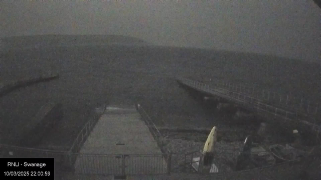 A dimly lit view of a pier extending over dark, choppy water. A sandy area is visible in the foreground, with a chain-link fence surrounding it. A yellow kayak is propped up near the fence, while other indistinct objects are scattered along the edge. In the background, the shoreline fades into the foggy atmosphere, and faint outlines of land can be seen in the distance. The overall scene conveys a desolate, overcast ambiance.