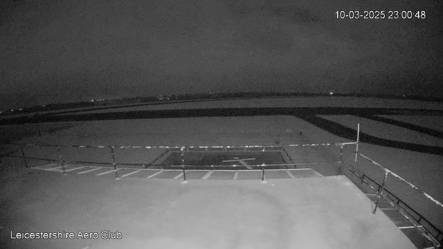 A black and white webcam image showing the nighttime view of an airport runway. The foreground features a flat surface with a landing pad marked, and a railing along the edge. In the background, there are faint lights visible in the distance. The overall scene is dark and lacks color detail. A timestamp in the corner indicates the image was captured on March 10, 2025, at 23:00:48.