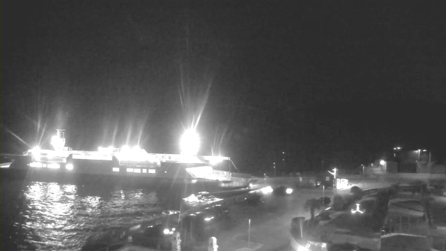 A dimly lit coastal scene at night featuring a large ferry illuminated by bright white lights. The ferry is partially docked at a port with reflective water surrounding it. In the foreground, there are outlines of vehicles and structures along the quay, along with additional indistinct lights in the background. The overall atmosphere is dark, with the ferry and its lights being the primary focus of the image.