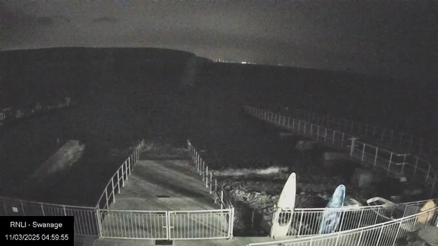 A dimly lit scene depicting a coastal area at night. In the foreground, there is a concrete walkway leading towards the water, surrounded by metal railings. On the left, two surfboards are positioned upright against the railing. The ocean is visible with faint waves lapping at the shore, and in the distant background, the silhouette of land can be seen. The sky is dark with some cloud coverage. A timestamp and location label are present in the bottom left corner.