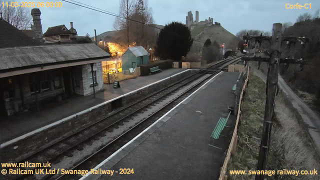 A railway station platform is depicted at dawn, with a light turquoise shed and benches on the left side. The station features a stone building with a thatched roof, while a wooden fence and tree create a border on the right. Railway tracks extend into the distance, leading to a hill topped with ruins partially obscured by trees in the background. The scene is illuminated by warm light from the shed, contrasted with the dim early morning sky.