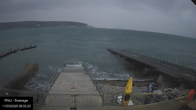 A cloudy scene of the coastline at Swanage, with a view of the dark blue sea meeting the shoreline. The water appears choppy, with waves splashing against a stone area. In the foreground, a set of steps lead down to the water, flanked by a railing. To the right, there are two boats on the shore: one is yellow and the other is blue. In the background, distant hills outline the coast, under a gray sky.