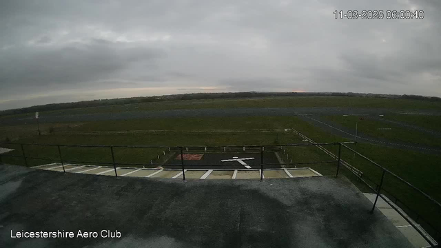 A view from a webcam at the Leicestershire Aero Club, showing a gray, overcast sky with minimal sunlight. Below, there is a grassy area with a large runway, and markings for a helipad that features a white cross on a dark surface. Surrounding the helipad are white boundary posts. The scene is quiet and open, with no vehicles or people visible.