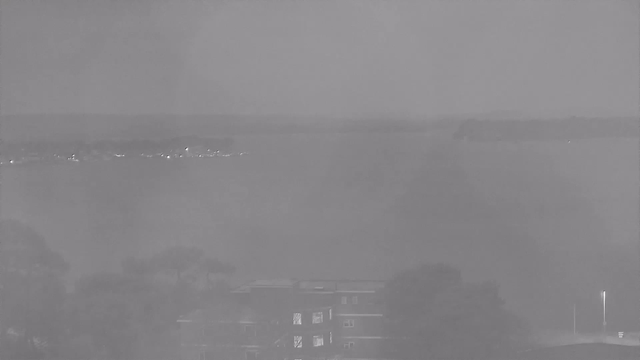 A monochromatic image showing a foggy landscape over a body of water. In the foreground, there are blurred outlines of trees and a building. The middle ground features a stretch of water, which fades into a misty horizon where faint lights from distant structures can be seen. The overall mood is hazy and subdued, with limited visibility.