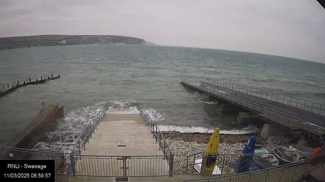 A cloudy sky hangs over a coastal scene with choppy waters. In the foreground, there are steps leading down to the water from a concrete walkway. To the left, a pier extends into the sea, partially submerged by waves. A rocky shore is visible to the right, alongside small boats with colorful hulls (yellow and blue). The background features a coastal cliff, indicating a hilly shoreline.