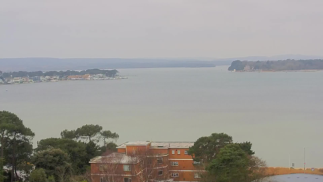 A view of a calm body of water surrounded by land. In the foreground, there are trees and a low-rise brick building. In the distance, a shoreline is visible with several boats and structures. The sky is overcast, creating a muted, grayish atmosphere. Farther away, rolling hills can be seen along the horizon.