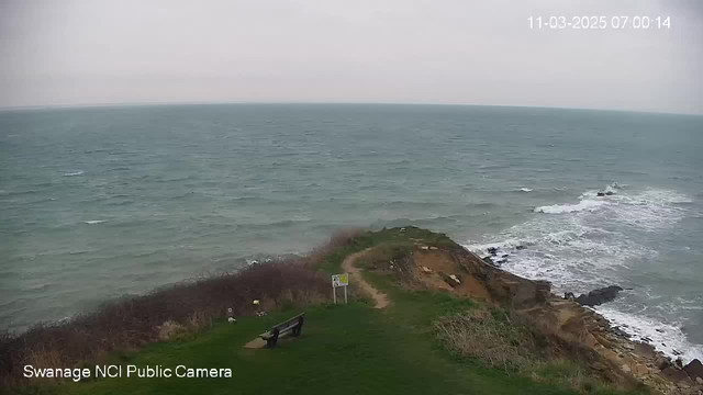 A coastal scene featuring a grey, overcast sky and choppy sea waters. In the foreground, there is a green grassy area with a bench facing the sea. To the right, rocky cliffs lead down to the surf. A warning sign is visible near the edge of the grass, providing caution about the area. The overall atmosphere is calm yet slightly turbulent, indicative of a breezy day by the ocean.