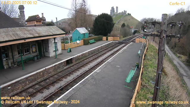 A train station scene featuring a stone building with a sloped roof on the left, housing benches and a poster. Green wooden benches are positioned along the platform. To the right, there are two sets of railway tracks leading away from the station. In the background, there's a hill with a castle ruin on top, surrounded by trees. A wooden fence encloses part of the station area, and a green sign indicating "WAY OUT" is visible near the platform edge. Above, a power pole with wires runs alongside the tracks. The sky is overcast.