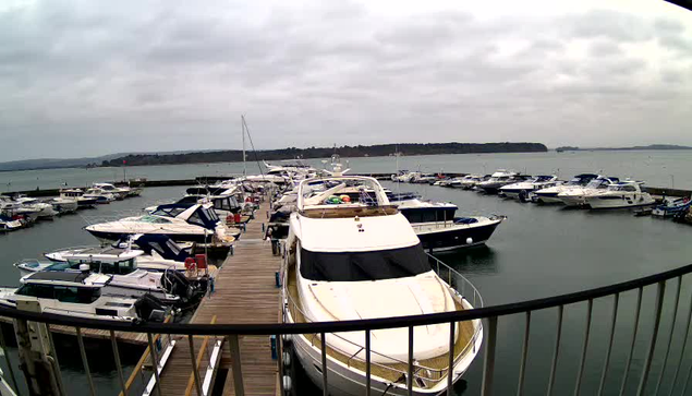 A marina scene under a cloudy sky, featuring multiple boats and yachts docked at the pier. The foreground shows a large white yacht with a covered seating area, while various smaller boats of different sizes and colors are moored alongside. To the right, additional yachts are lined up with a view of water extending towards the horizon, where land is faintly visible in the distance. The wooden dock is visible, leading into the marina area.