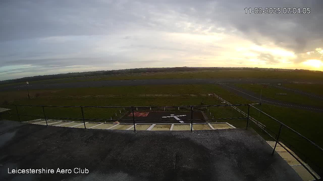 A cloudy sky is visible with light patches indicating the rising sun. The foreground features a concrete platform with a railing, looking out over a grassy airfield. A white cross is marked on the ground, indicating a helipad or landing area, with a few scattered objects visible on the field. The scene is peaceful, suggesting early morning at the Leicestershire Aero Club.