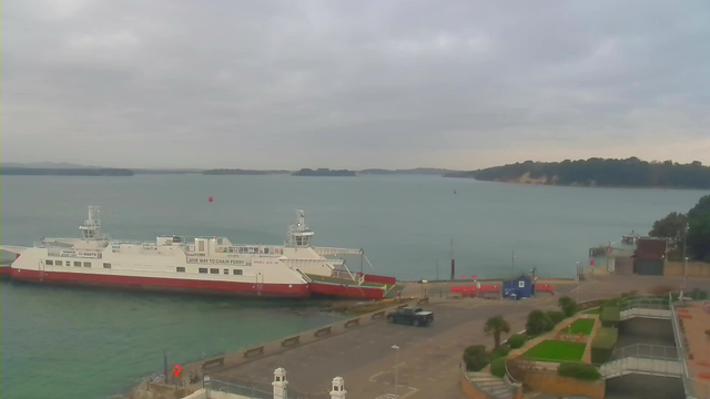 A ferry is docked at a port, with a view of calm waters and distant islands. The sky is cloudy, and there are several boats in the water. Near the dock, there is a red buoy and a small blue building. The area is surrounded by greenery and pathways, with some palm trees visible. A vehicle is parked nearby on the promenade.