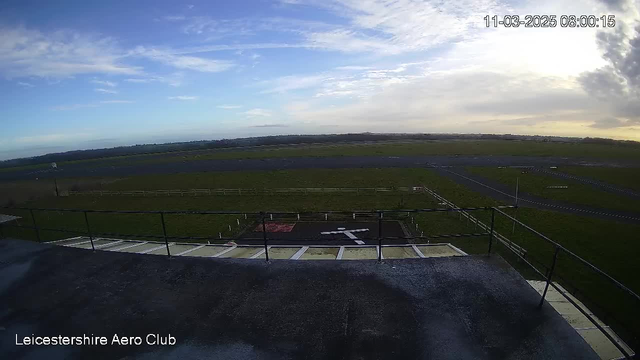 A wide view of an aviation area showing a clear sky with some clouds and an expansive grassy field. In the foreground, there is a railing and a flat rooftop surface. Below, a helipad is marked with a white cross on a dark background. In the background, there are runways and a fenced area, indicating an airfield setting. The scene captures a tranquil morning atmosphere.