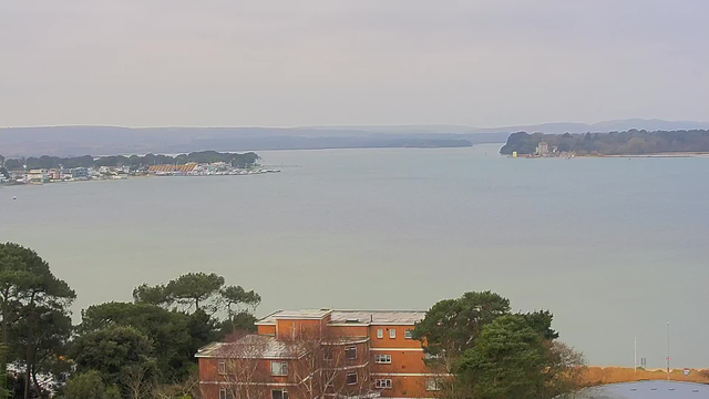 A view of a calm body of water, with a shoreline on the left featuring buildings and trees. In the background, there are additional buildings along the coast and hills in the distance, under a cloudy sky. There are some boats visible on the water, and the landscape is predominantly green and gray. A brick building is in the foreground on the right side of the image, surrounded by trees.