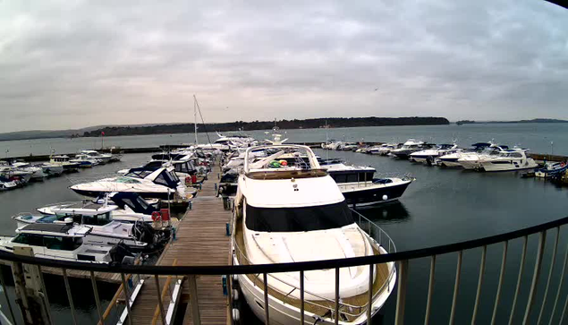 A view of a marina on a cloudy day, featuring numerous parked boats and yachts of various sizes. The boats are docked along wooden piers, with calm water reflecting the overcast sky. In the background, there are distant green hills and a few trees, adding to the serene atmosphere of the waterfront. The scene conveys a peaceful, maritime setting.