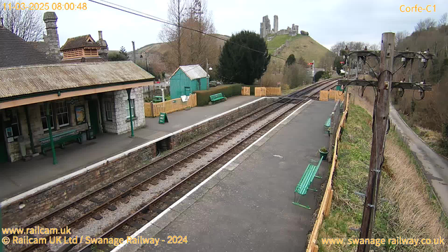 A view of Corfe Castle railway station featuring a stone building with a sloped roof and several green benches along the platform. In the background, there are rolling hills with the ruins of Corfe Castle on top, surrounded by bare trees. A nearby green shed and a section of wooden fencing frame the area. The scene is mostly clear with a gray sky. The railway tracks run parallel to the platform, leading into the distance.