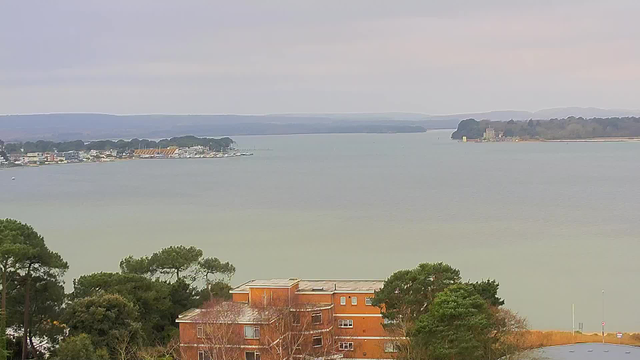 A view of a calm bay with water reflecting a cloudy sky. In the foreground, there is a building with a reddish exterior and several trees with green foliage. Across the bay, there are scattered homes and boats, along with a stretch of land lined with trees. The horizon features distant hills.