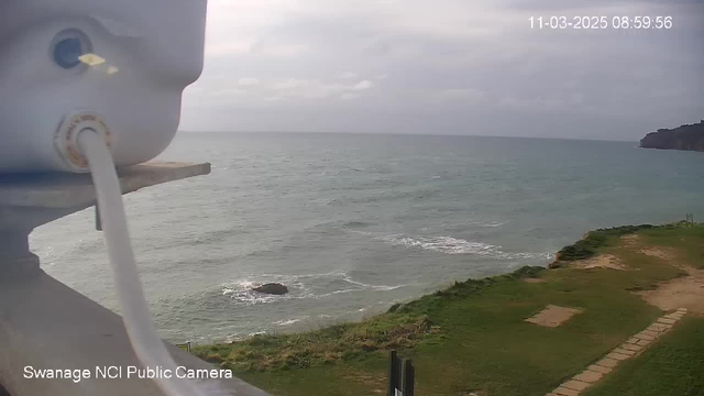 A view from a webcam showing a coastal scene. In the foreground, there is a white device attached to a railing, with a cable extending from it. Beyond, the ocean is visible with gentle waves lapping at the shore. The sky is overcast with gray clouds, and patches of grass and a pathway can be seen on the land next to the water. A large rock is partially submerged in the water near the shoreline. The image has a timestamp indicating the date and time.