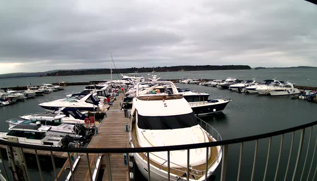 A view from a webcam showing a marina filled with various yachts and boats. The foreground features a large white yacht with a prominent bow. Several smaller boats, some in blue and others in white, are moored along a wooden dock. In the background, the water is calm, and a cloudy sky is visible, with hints of land in the distance. The overall atmosphere is tranquil and maritime.