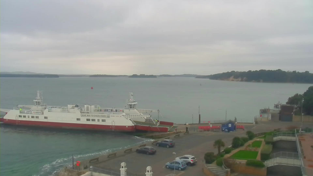 A cloudy scene of a body of water with a ferry docked at a terminal. The ferry is mostly white with red accents and has a sign indicating a speed limit. There are several parked cars in the foreground, and a small blue building and red container near the dock. In the background, hills and trees outline the horizon. The water appears calm with gentle ripples.