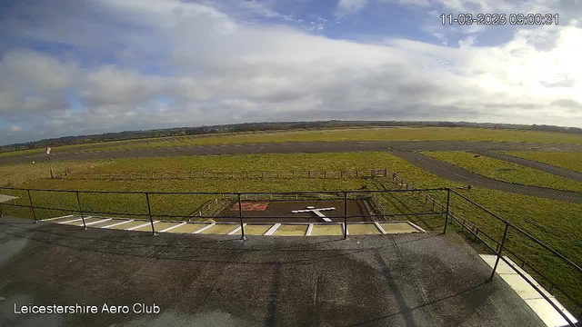 A view from a webcam at the Leicestershire Aero Club showing a wide, open landscape. In the foreground, there is a railing along the edge of a building or platform. Below, a grass area features a large "X" marking in white paint, possibly indicating a helicopter landing zone. The background consists of a grassy airfield bordered by wooden fencing, with several small roads visible. The sky is partly cloudy, and the scene is bright with daylight. The date and time at the top right indicate it is March 11, 2025, at 9:00 AM.