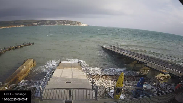 A view of a seaside area with gray skies and turbulent water. In the foreground, there are steps leading down to the water, flanked by a metal railing. To the left, a wooden pier extends into the sea, while a wider pier is visible on the right side of the image. Along the shore, there are kayaks stored, including a yellow one. The rocky beach is visible beneath the water's edge, with waves crashing against the steps and shoreline.