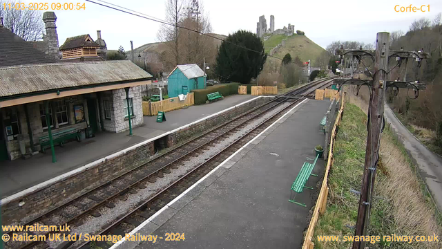 A view of Corfe Castle railway station on a cloudy day, with the ruins of Corfe Castle visible on a hill in the background. The station platform features a covered waiting area with green benches and posters. There is a green "WAY OUT" sign visible on the platform. Railway tracks extend to the right, while a wooden telephone pole stands on the edge of the platform. Surrounding vegetation includes trees and a grassy area.