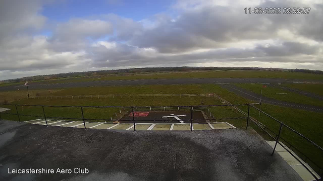 A view from a webcam at Leicestershire Aero Club showing an expansive landscape. In the foreground, a flat rooftop with a railing overlooks an airstrip. The airstrip is paved and bordered by green grass and patches of dirt. A helipad marked by a white cross is visible to the left of the image. The skies above are mostly cloudy, with some blue patches visible. The scene conveys a calm and open atmosphere typical of an airfield.