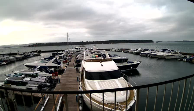 A marina scene showing multiple boats docked in a calm harbor. The foreground features a large white yacht with a rounded top. In the background, numerous smaller boats and yachts are moored along the wooden pier. The sky is overcast with gray clouds, and the water reflects the cloudy sky. The shoreline is visible in the distance with hills or trees lining the horizon.