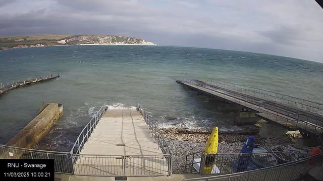 A view of a coastal area featuring a ramp leading down to a rocky beach, with the ocean waves gently lapping at the shore. In the foreground, there are yellow and blue kayaks positioned against a railing. In the background, cliffs rise in the distance under a partly cloudy sky. The image captures a tranquil seaside scene with calm waters.