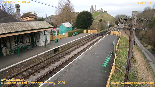 A view of Corfe Castle railway station showing two railway tracks, a waiting area with green benches, and a stone building with a sloped roof. In the background, Corfe Castle is visible on a hilltop, surrounded by trees. There's a wooden fence along the platform, with a sign that reads "WAY OUT." Electrical poles are present beside the tracks, and the scene is set under a cloudy sky.