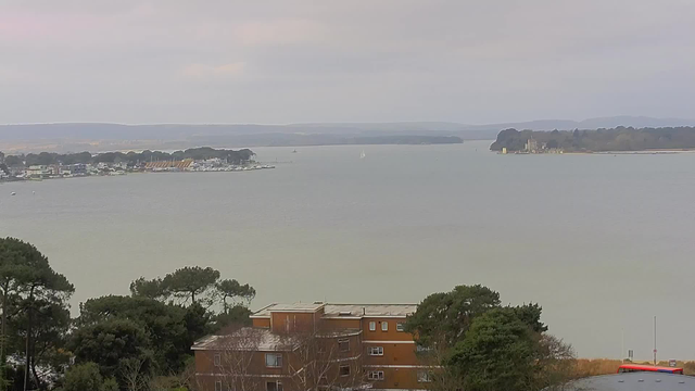 A view of a calm body of water during overcast weather. In the foreground, there are trees and low buildings, including a reddish-brown structure. The water stretches out towards the background, where a series of boats and a small sailboat can be seen. On the far side of the water, there are trees and a landmass with a building that resembles a castle. The atmosphere appears tranquil and slightly hazy, with a soft gradient in the sky.