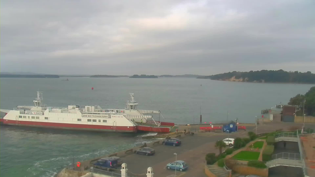 A white and red ferry is docked at a harbor. The water is calm with several small boats in the distance. In the foreground, there are parked cars on a paved area, with some greenery and low hedges visible. The sky is overcast, creating a muted light.