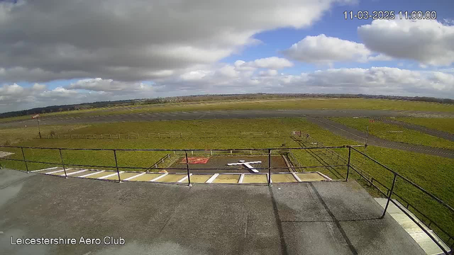 A view from a high vantage point overlooking an airfield. The foreground features a gray, flat rooftop with a railing. Below, there is a grassy area divided by a dirt runway, with a white cross marked on the ground. The background shows a wide expanse of grass and a cloudy sky, with patches of blue peeking through. There are few trees in the distance, and the landscape appears open and expansive.