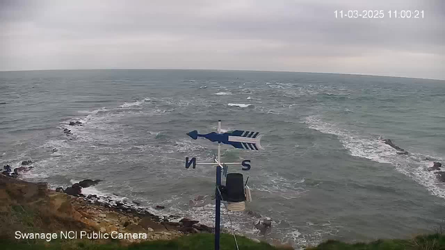 A view of the ocean with waves gently rolling against a rocky shore. In the foreground, there is a weather vane marked with the directions "N" (north) and "S" (south). The sky is overcast with gray clouds, and the scene appears tranquil but slightly windy. The image is timestamped in the corner, indicating the date and time.